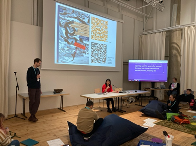 A symposium with person standing to the left holding a microphone; another in the middle, seated, in front of a projection with book cover and QR codes and next to a screen showing live captioning; more people in the foreground on different forms of seating and being;
