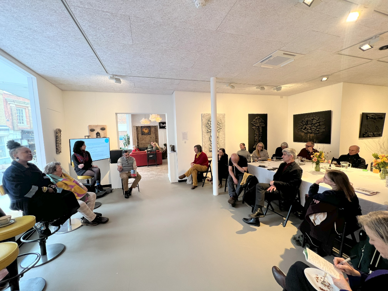 A group of people seated around a table on the right of the photos in a room painted white watching a panel of people seated on high stools on the left.