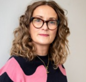 A headshot of a woman with curly shoulder length hair, wearing glasses and a pink and black jumper.